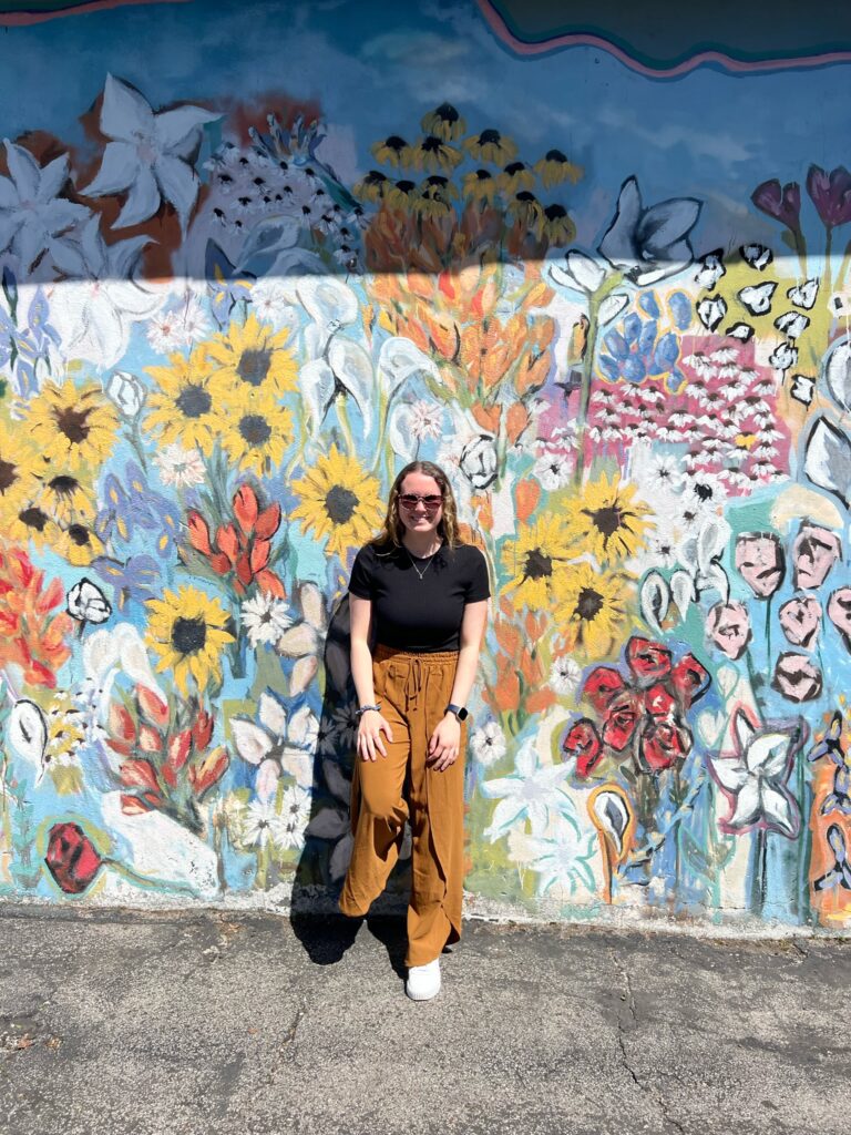 Liv, food freedom dietitian, posing in front of a colorful, flower mural wearing orange flowy pants and a back tshirt